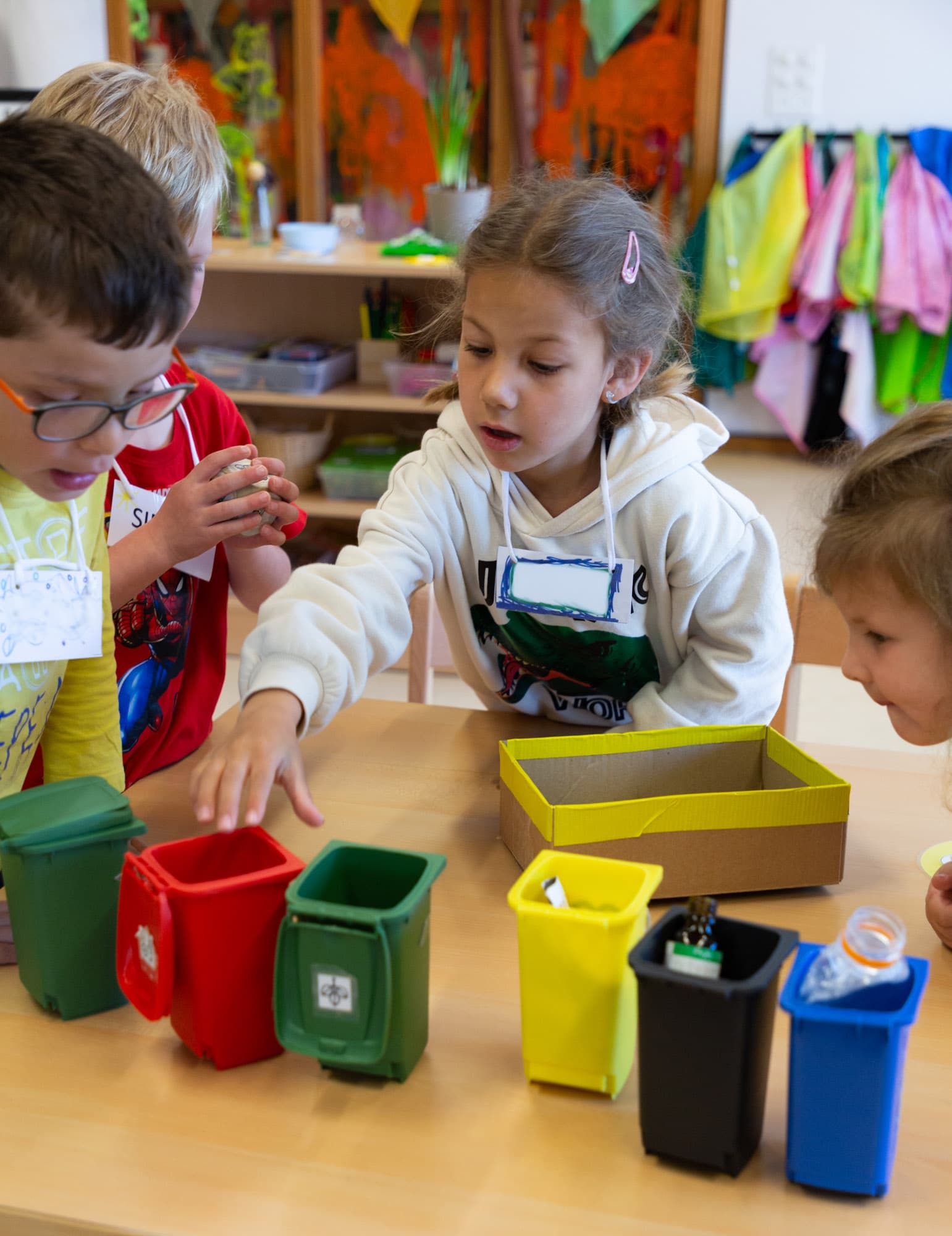 Zwei Mädchen und zwei Jungs sitzen um einen Tisch im Kindergarten, auf dem verschiedene kleine Plastikkübel symbolisch für Recyclingcontainer stehen.