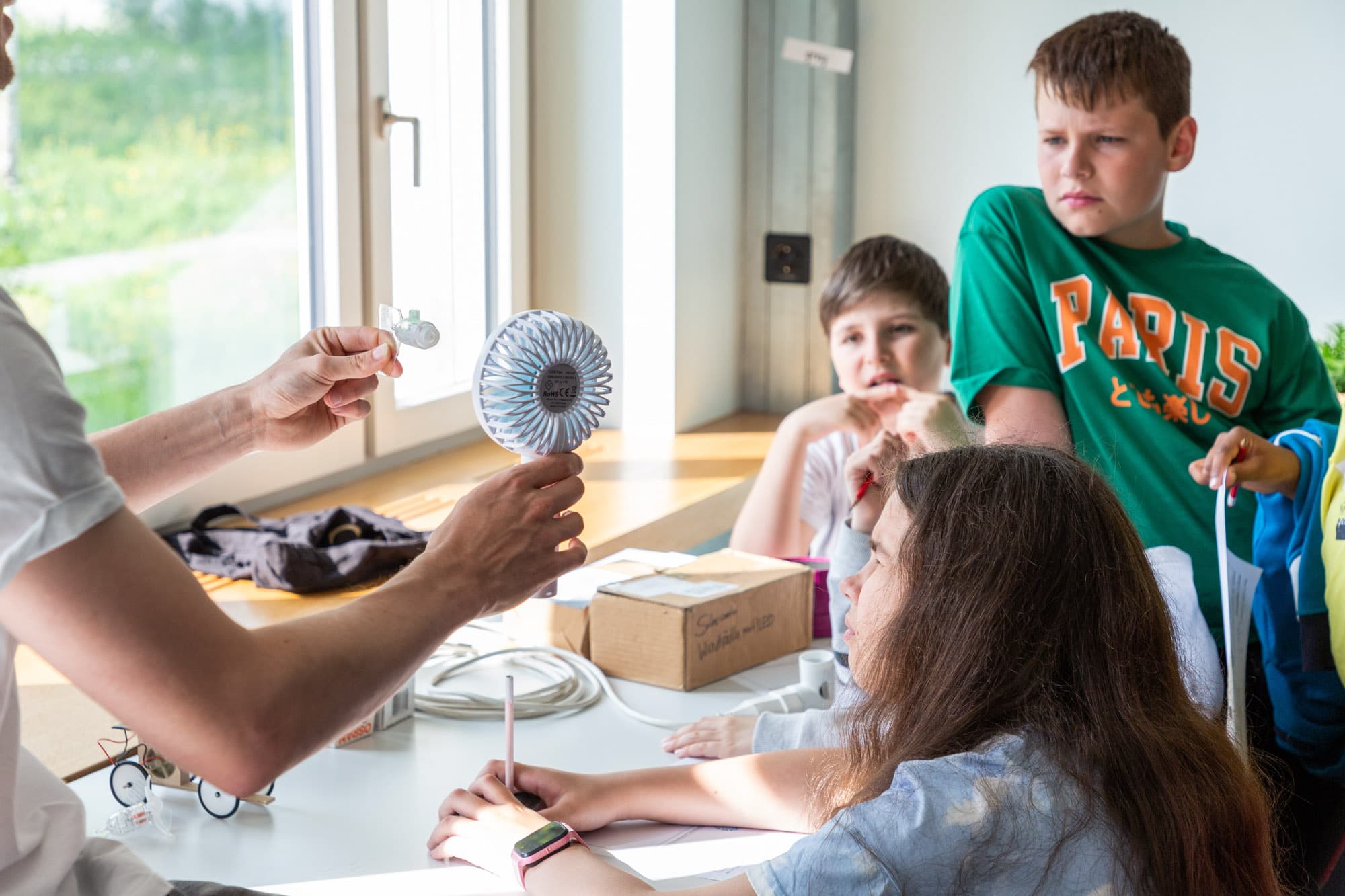 Kinder schauen einem Erwachsenen zu, wie er mit einem kleinen Ventilator die Windkraft symbolisch zeigt.