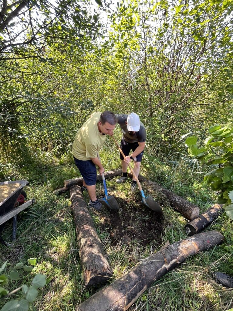 Zwei Männer schaufeln innerhalb einer Totholzstruktur aus vier Stämmen.